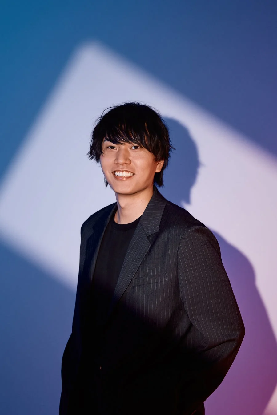 青と紫の光が当たる背景の前で、黒いストライプのスーツを着た若い男性が笑顔でポーズをとっているポートレート。